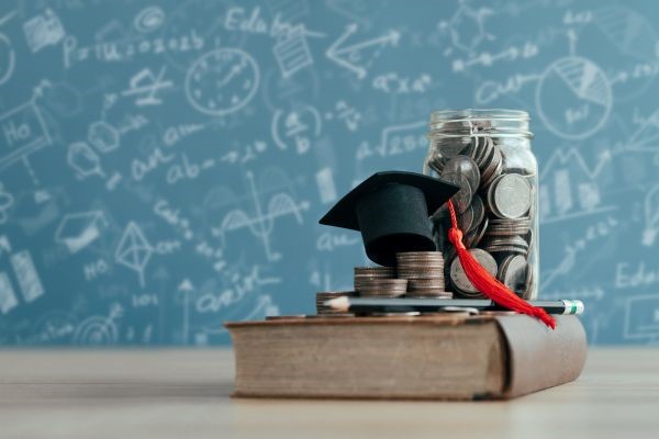 A graduation cap and a jar of money sitting on a book in front of a chalkboard