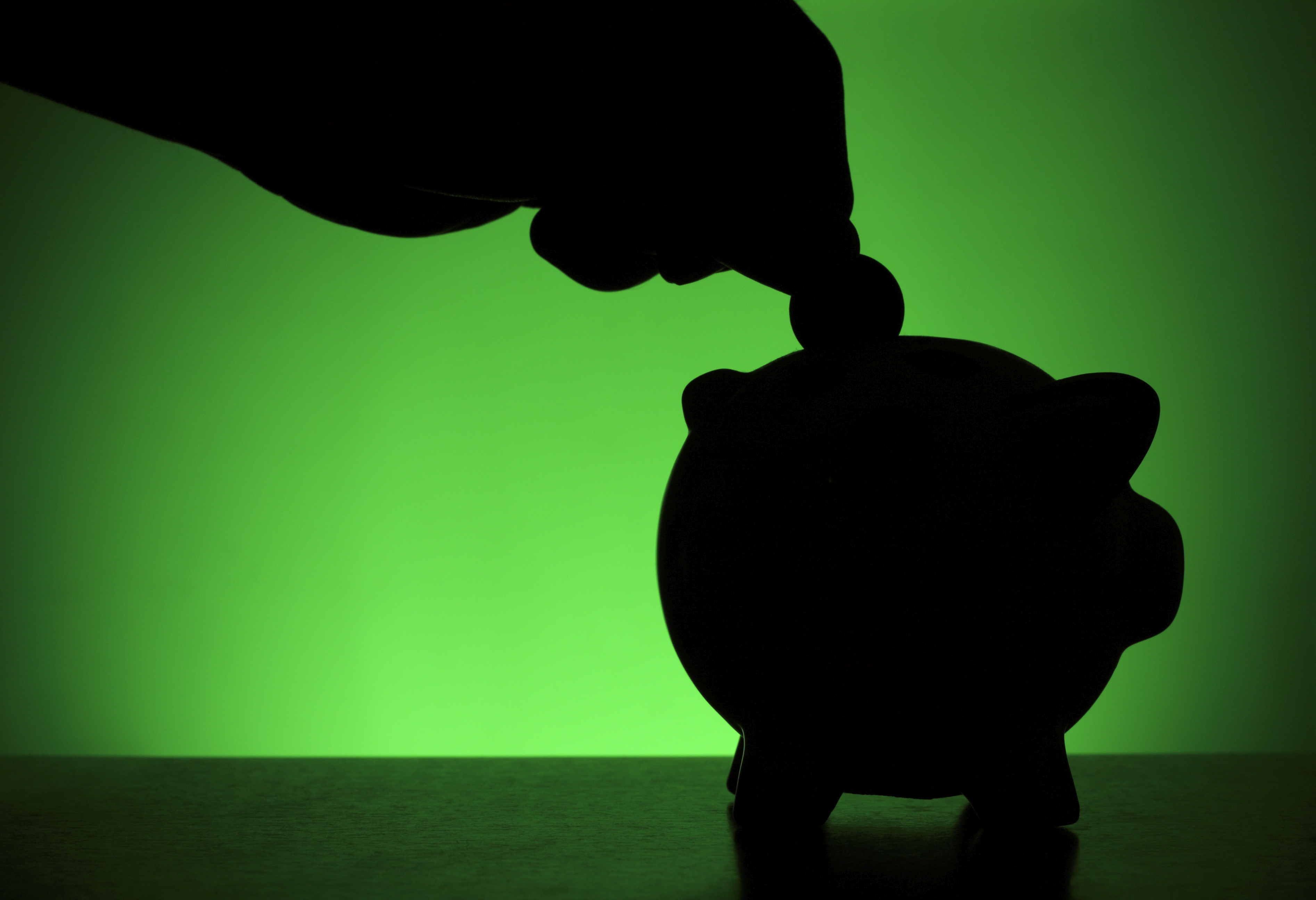 silhouette of a hand placing a coin into a piggy bank set against a green background