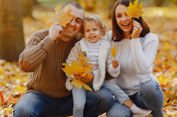 Mom and Dad sit in a pile of leaves with young daughter